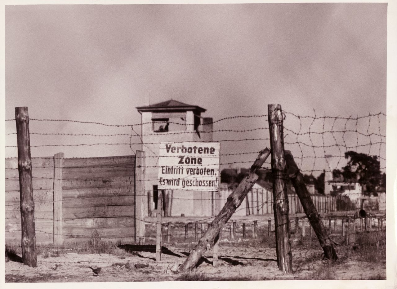 Schwarz-weiß Foto von der Einzäunung des Internierungslagers und dem Schild: Verbotene Zone. Eintritt verboten. Es wird geschossen!, Im Hintergrund ist ein Wachturm zu sehen. Rückseite: handschriftlich: Copyright by Perlia.