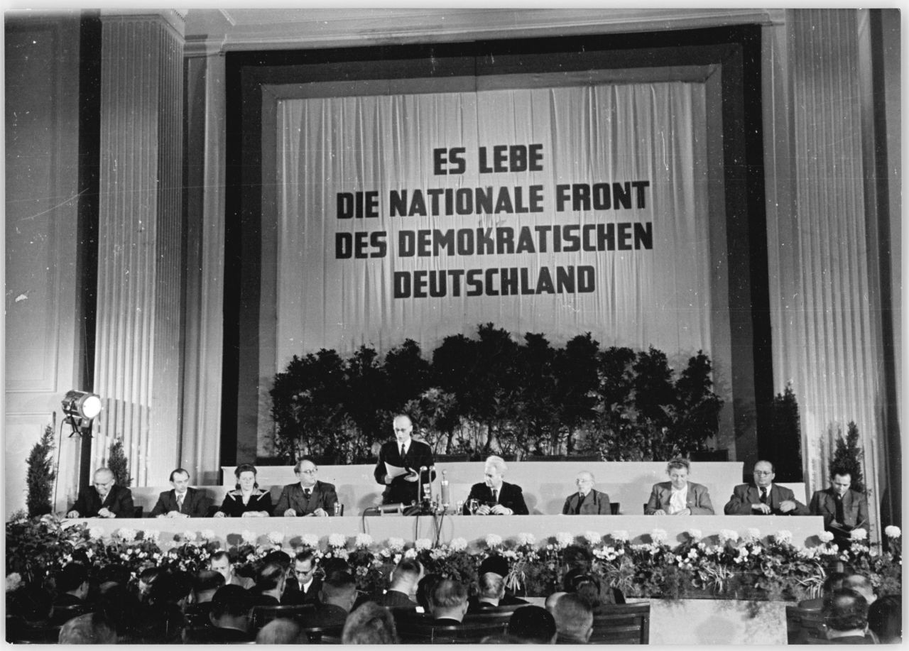 Schwarz-weiß-Fotografie der konstituierenden Sitzung der Provisorischen Volkskammer der DDR. Auf einem Vorhang hinter dem Podest ist in Großbuchstaben zu lesen: Es lebe die nationale Front des demokratischen Deutschland